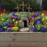Wooden raised garden bed with resurrection garden display featuring three crosses on hill, stone tomb, and colorful spring flowers